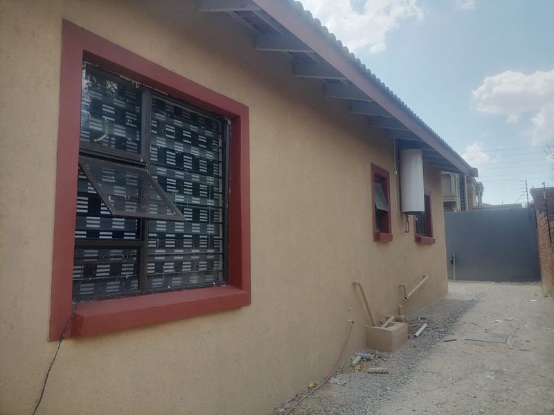 Rear exterior view of the apartment showing the building facade, external geyser, steel window frames with burglar bars, and unfinished ground area.