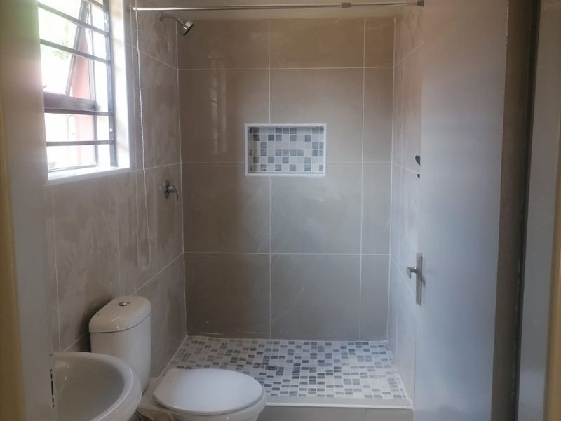 A clean, functional bathroom featuring a walk-in shower with a mosaic-tiled niche, pedestal basin, and toilet, finished with neutral wall and floor tiles.