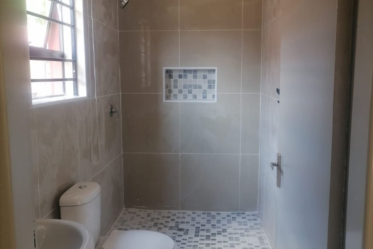 A clean, functional bathroom featuring a walk-in shower with a mosaic-tiled niche, pedestal basin, and toilet, finished with neutral wall and floor tiles.