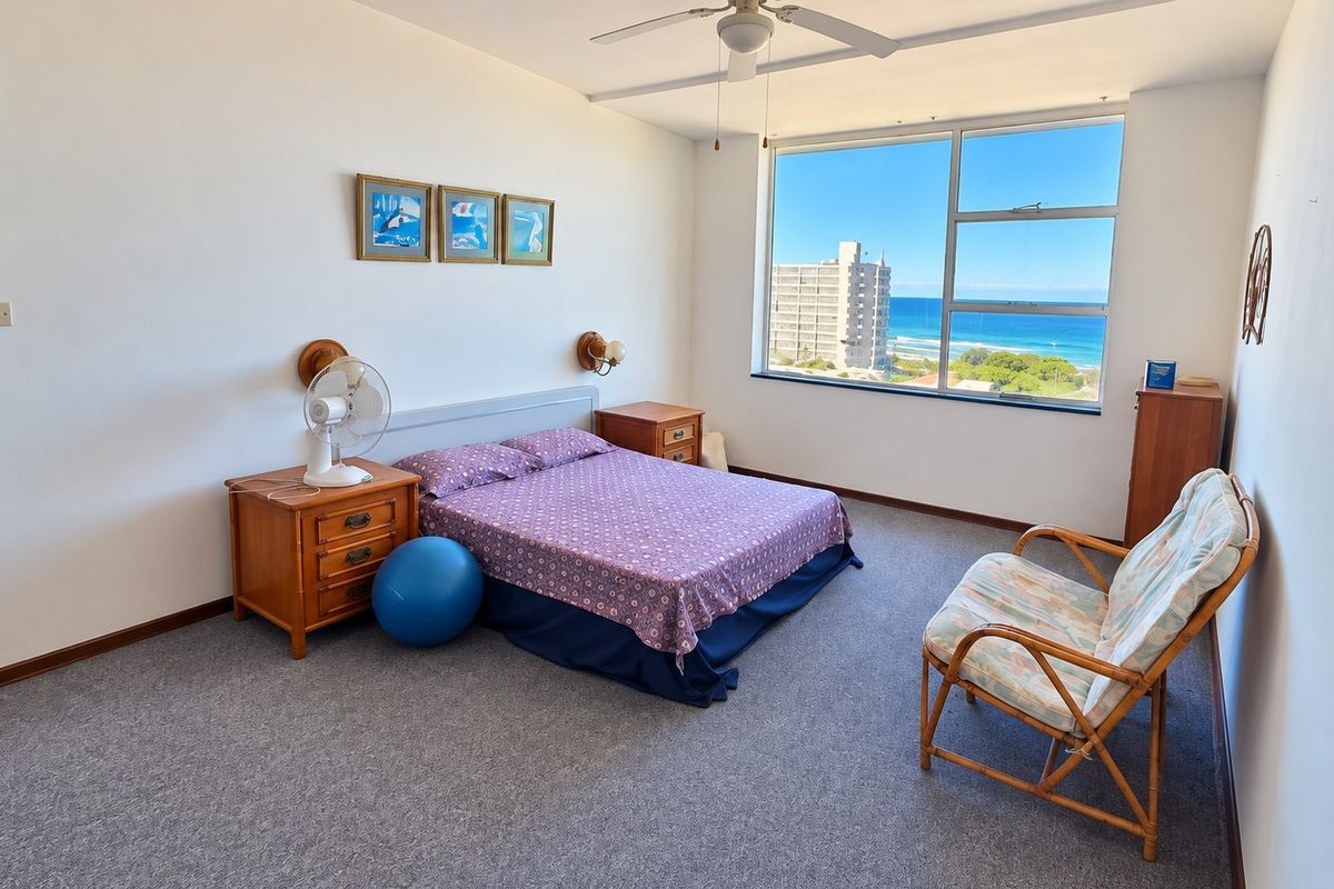 A spacious bedroom featuring carpet flooring, a ceiling fan, and a large window offering scenic ocean and coastal views.