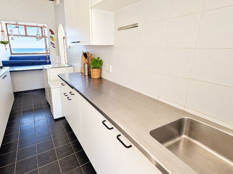 A functional galley-style kitchen featuring stainless steel countertops, a built-in sink, tiled backsplash, and white cabinetry, with a view towards the living area.