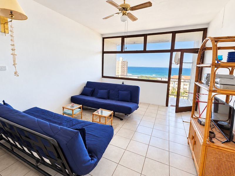 A bright living room featuring tiled flooring, a ceiling fan, and large windows providing expansive ocean views and access to a balcony.