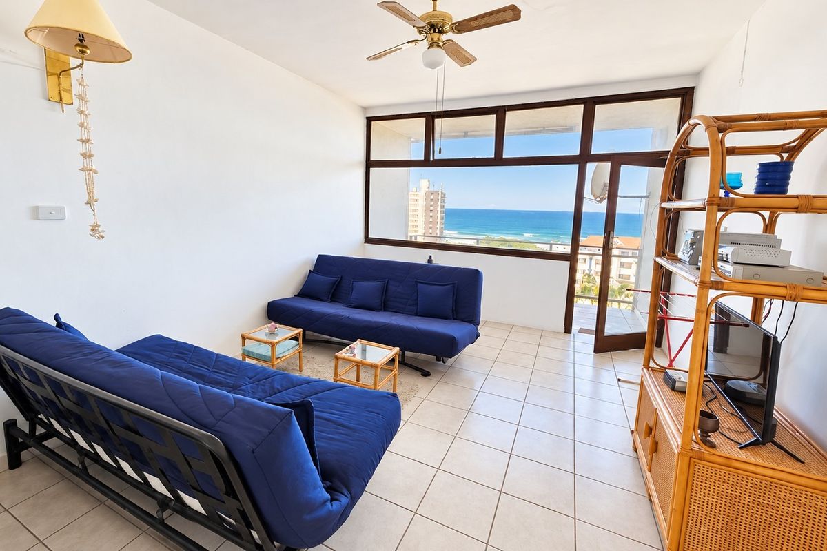 A bright living room featuring tiled flooring, a ceiling fan, and large windows providing expansive ocean views and access to a balcony.