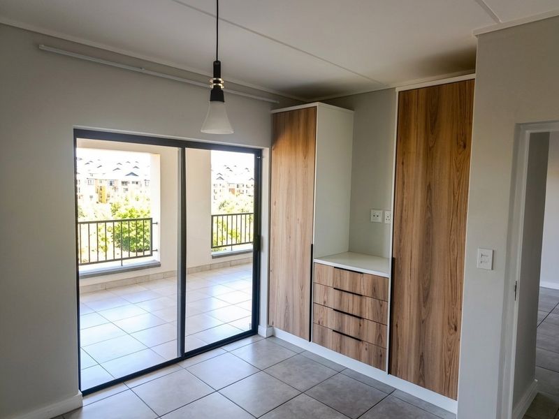 A bright bedroom featuring modern built-in cupboards with wood-grain finish, tiled flooring, and a sliding glass door leading to a private balcony.