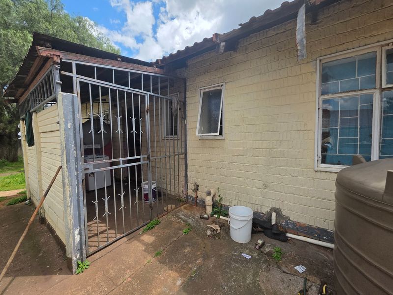 Rear exterior view of the property showing a paved area, security gate, and brick wall with visible signs of wear and maintenance requirements.