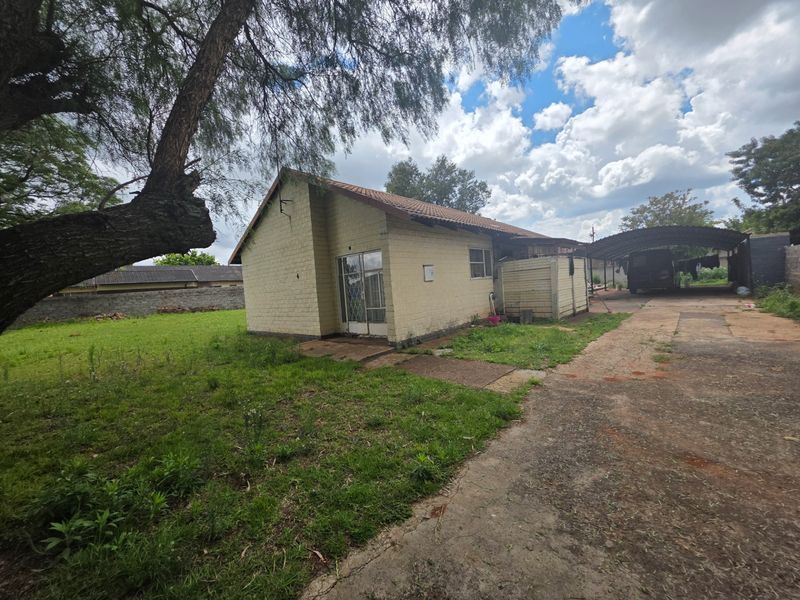 Rear view of the property featuring a brick exterior, tiled roof, concrete driveway, and a carport structure set on a large grassed lot.