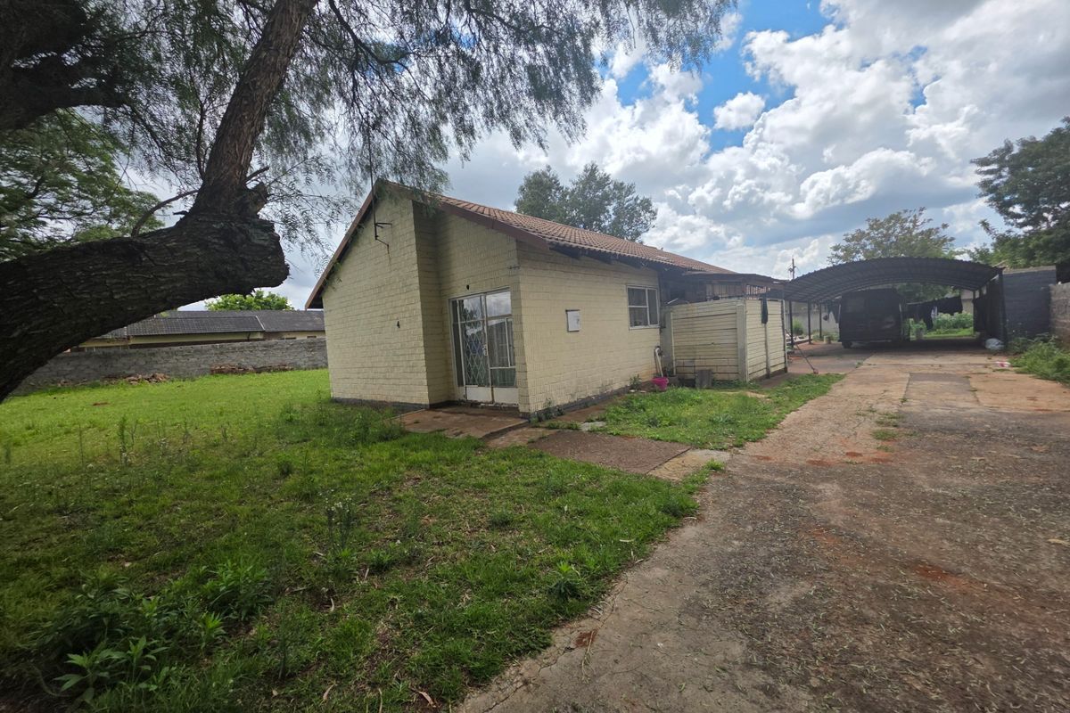 Rear view of the property featuring a brick exterior, tiled roof, concrete driveway, and a carport structure set on a large grassed lot.