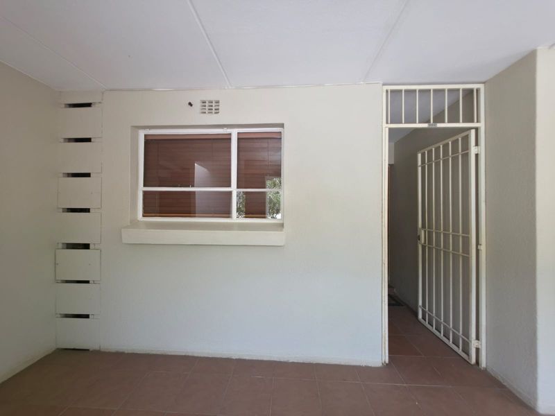 A covered patio area featuring tiled flooring, a secure entrance gate, and a window with burglar bars, providing a functional outdoor transition space.