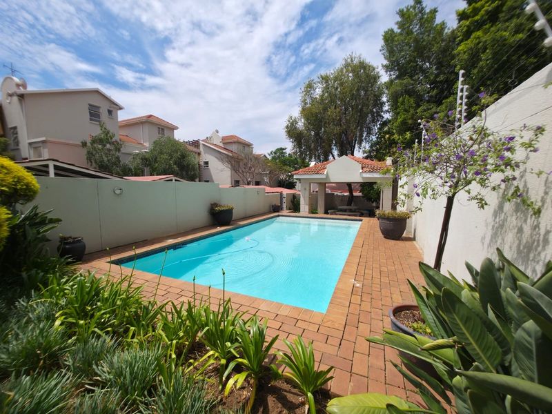 A well-maintained outdoor pool area featuring a rectangular swimming pool, brick paved surround, and a covered patio structure within a secure walled garden.