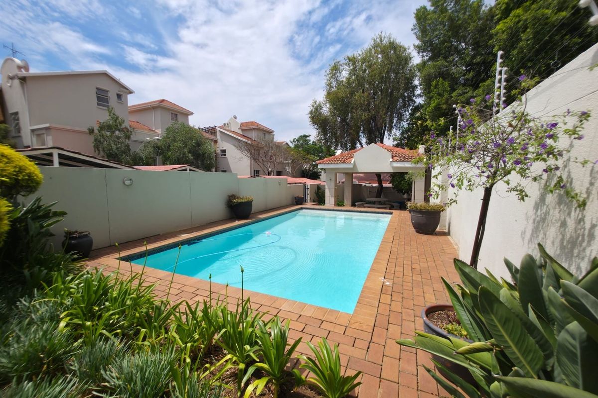 A well-maintained outdoor pool area featuring a rectangular swimming pool, brick paved surround, and a covered patio structure within a secure walled garden.