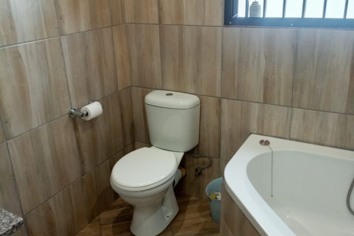A compact bathroom featuring wood-look ceramic wall and floor tiles, a standard toilet, and a built-in bathtub with security bars on the window.