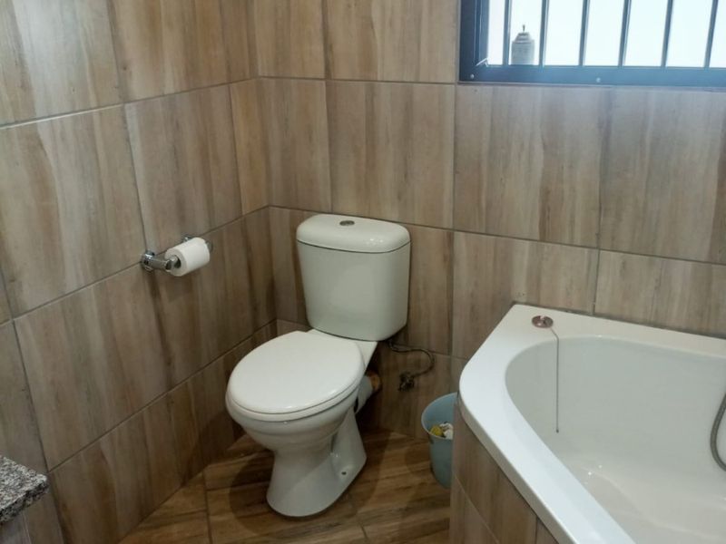 A compact bathroom featuring wood-look ceramic wall and floor tiles, a standard toilet, and a built-in bathtub with security bars on the window.