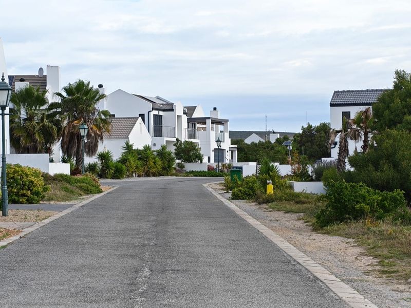 A view of a paved residential street within a housing estate, featuring well-maintained white architectural facades and landscaped surroundings.