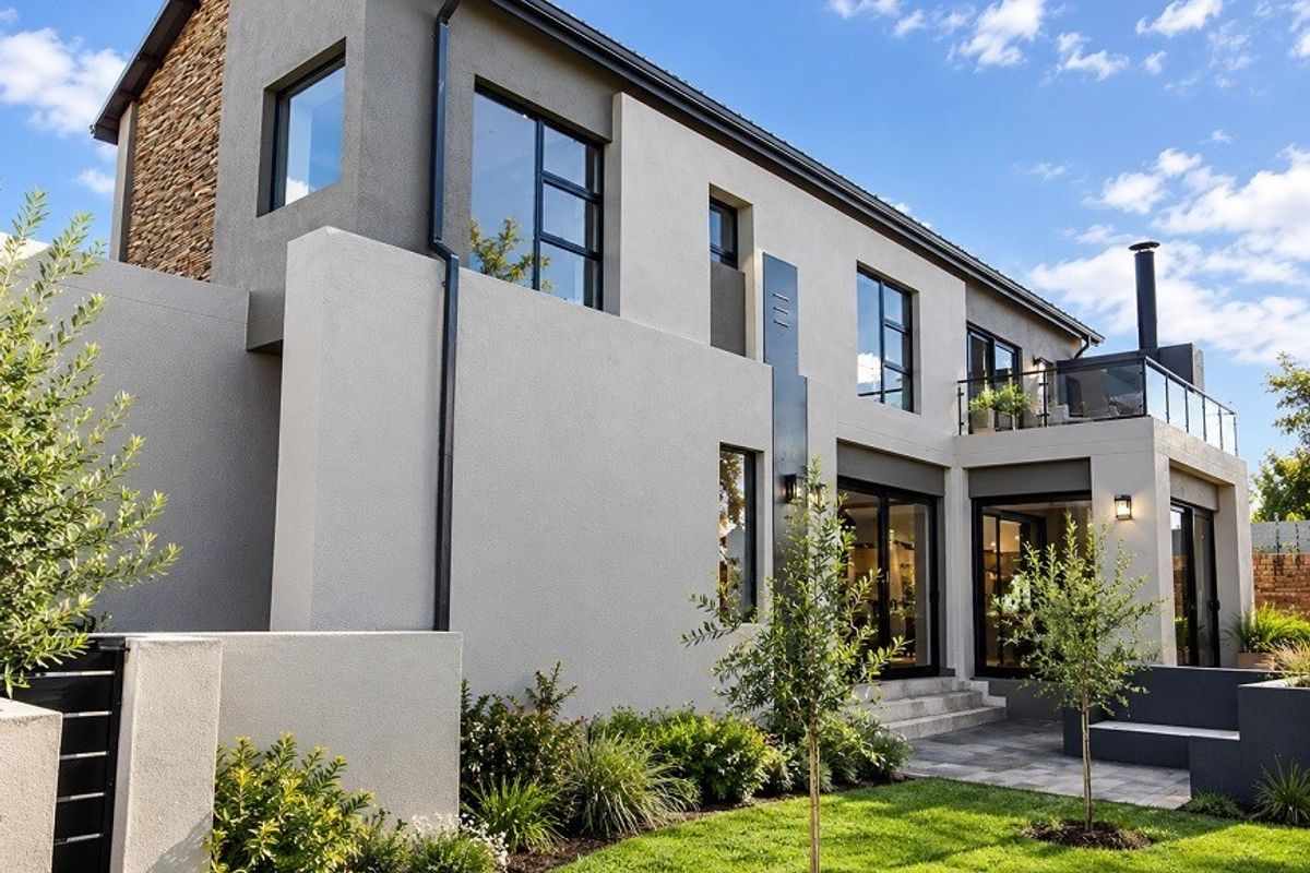 Modern double-storey rear elevation featuring stone cladding, a spacious balcony with glass balustrade, paved patio area, and well-maintained landscaped garden.