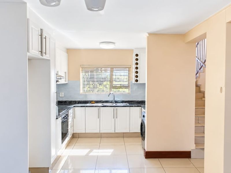 A clean, functional kitchen featuring white cabinetry, dark stone countertops, built-in appliances, and tiled flooring, with an adjacent staircase leading to the upper level.
