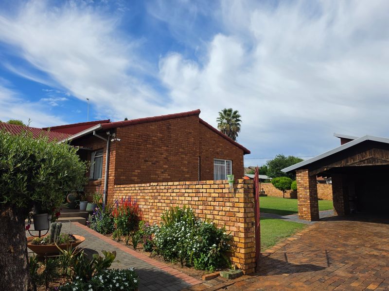 A well-maintained rear exterior view featuring a face brick house, a paved driveway, a carport, and a landscaped garden with a boundary wall.