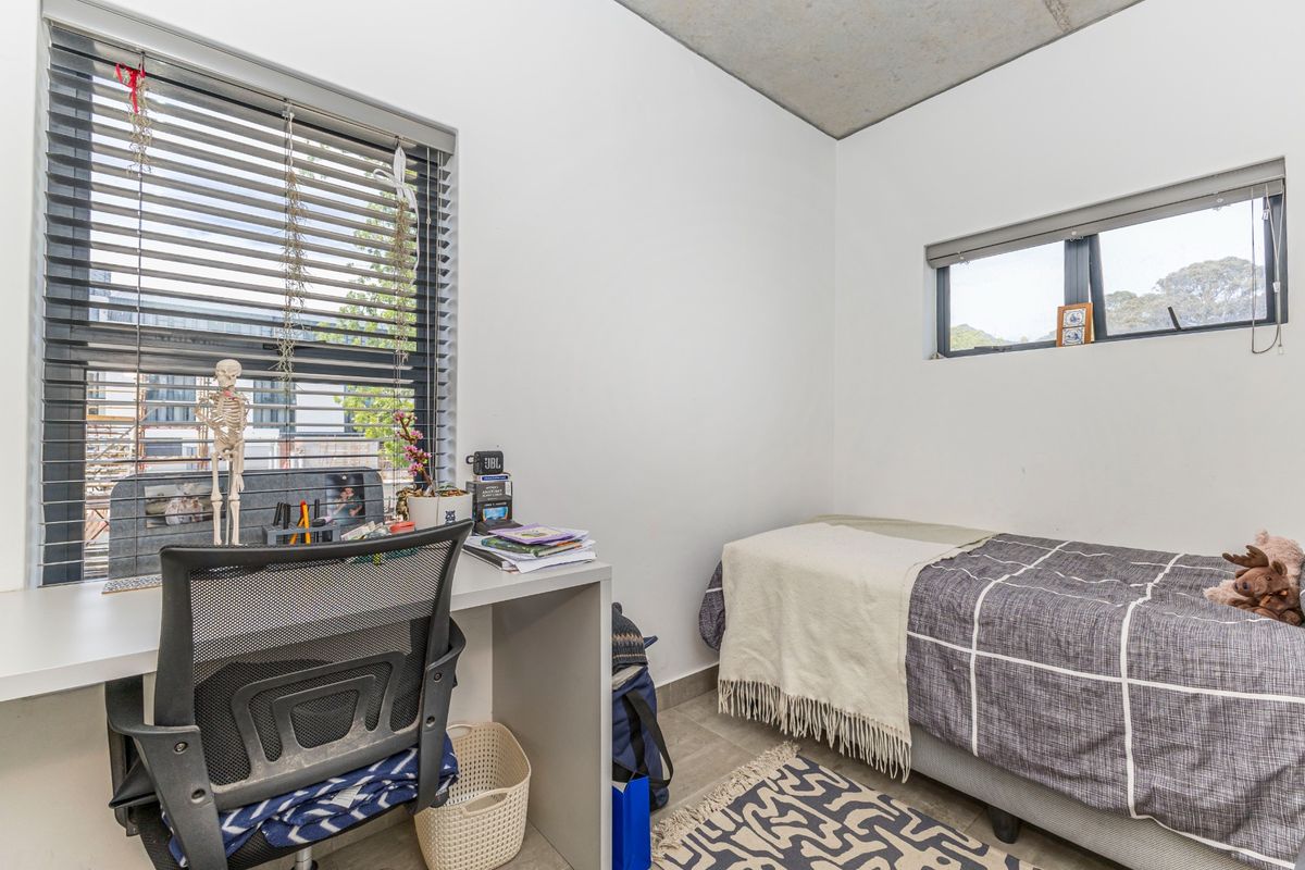 A compact bedroom featuring a built-in desk, exposed concrete ceiling, tiled flooring, and large windows with venetian blinds.