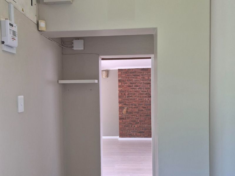 A neutral hallway featuring laminate flooring and coving, leading into an adjacent room with an exposed brick feature wall.