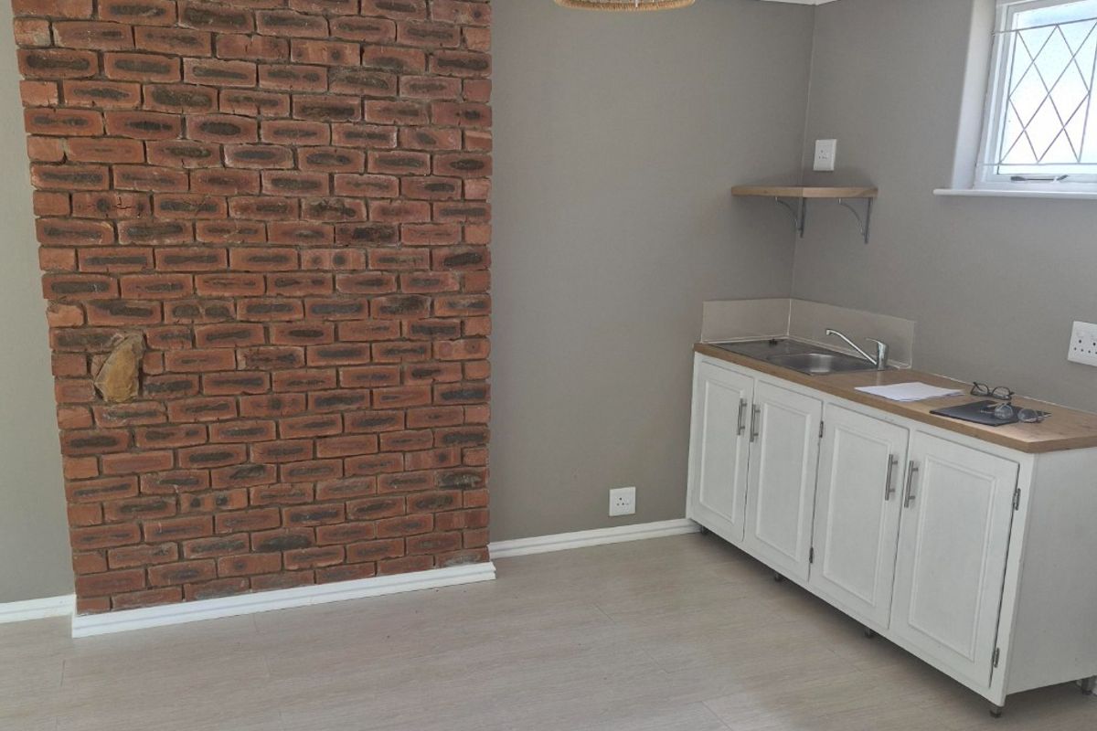 A compact kitchen area featuring a rustic exposed brick feature wall, white cabinetry with a stainless steel sink, and light-toned laminate flooring.