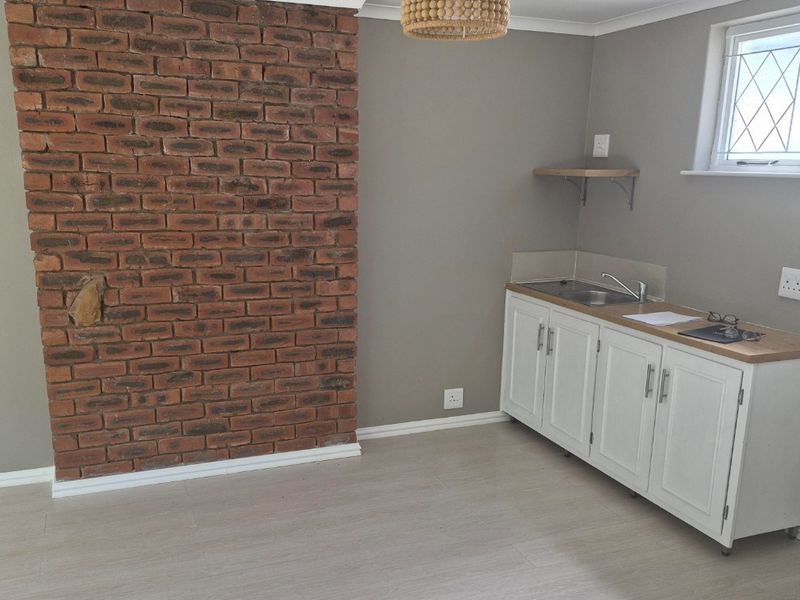 A compact kitchen area featuring a rustic exposed brick feature wall, white cabinetry with a stainless steel sink, and light-toned laminate flooring.