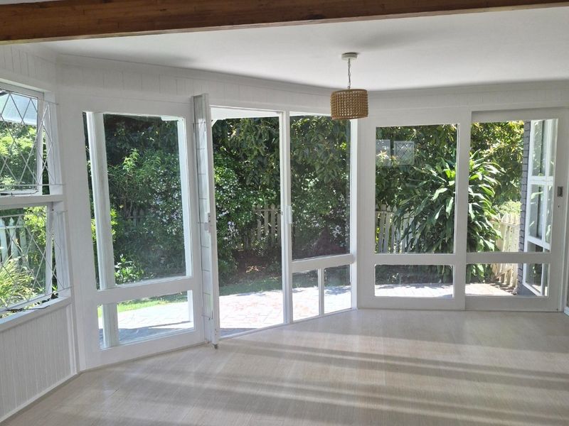 Bright and airy living room featuring large glass windows and doors, exposed timber ceiling beam, and light-toned laminate flooring with direct garden access.