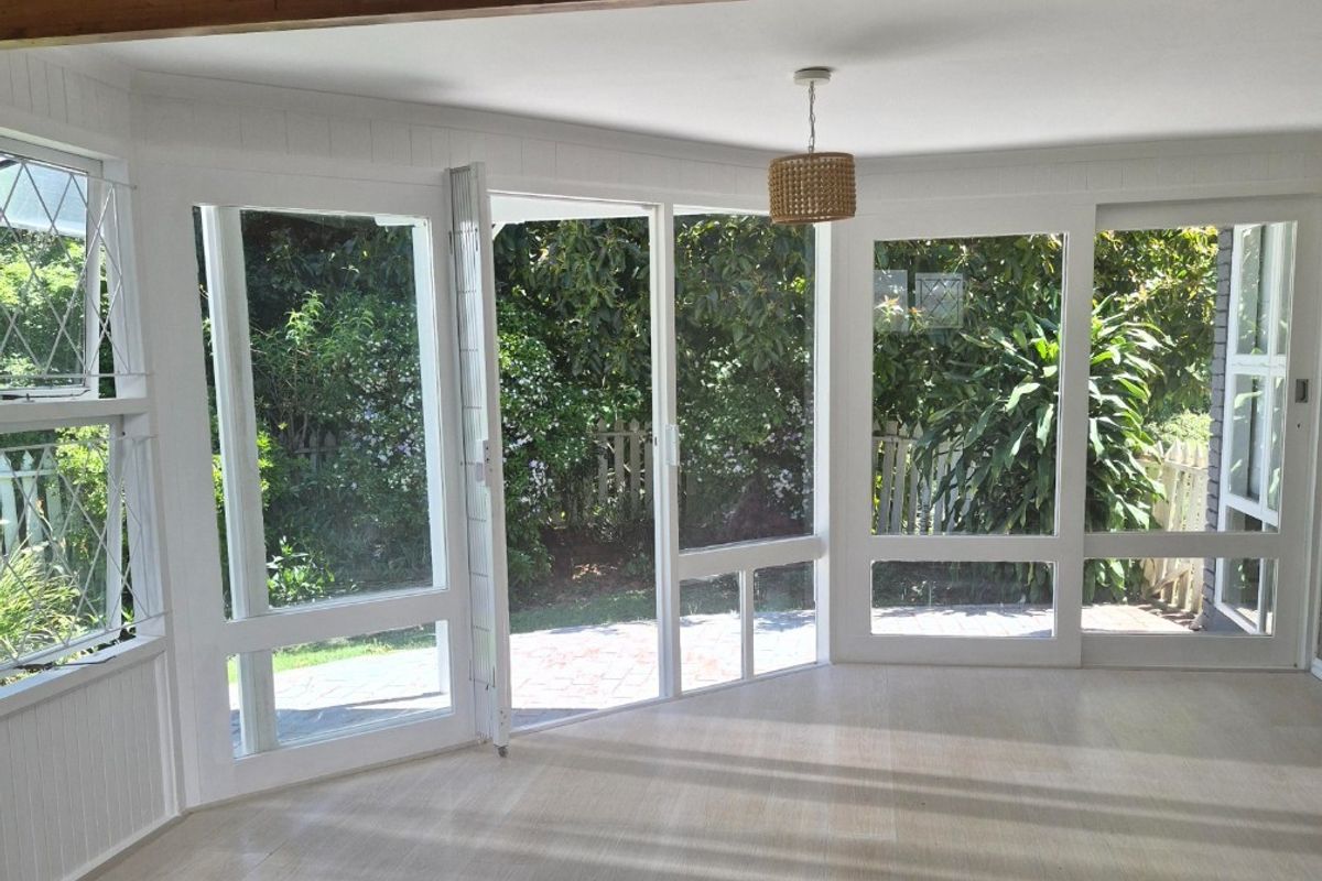 Bright and airy living room featuring large glass windows and doors, exposed timber ceiling beam, and light-toned laminate flooring with direct garden access.