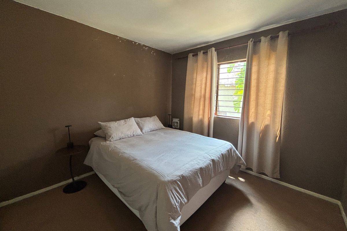 A bedroom featuring carpeted flooring, security bars on the window, and a ceiling light fixture, currently showing signs of wall surface wear.