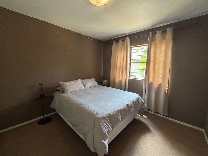 A bedroom featuring carpeted flooring, security bars on the window, and a ceiling light fixture, currently showing signs of wall surface wear.