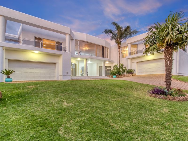 A modern, double-story architectural home featuring a spacious paved driveway, double garages, and expansive balconies with glass balustrades set in a manicured garden.
