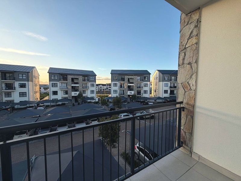 A private balcony featuring a metal balustrade and stone-clad pillar, offering an elevated view over the apartment complex parking area.