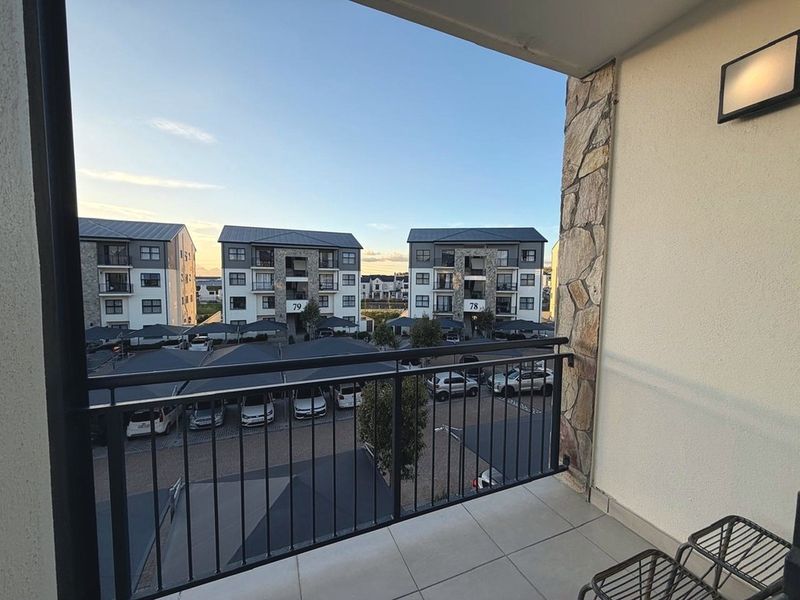 A clean, modern balcony featuring tiled flooring, stone cladding accents, and a metal balustrade, offering an elevated view of the apartment complex parking area.