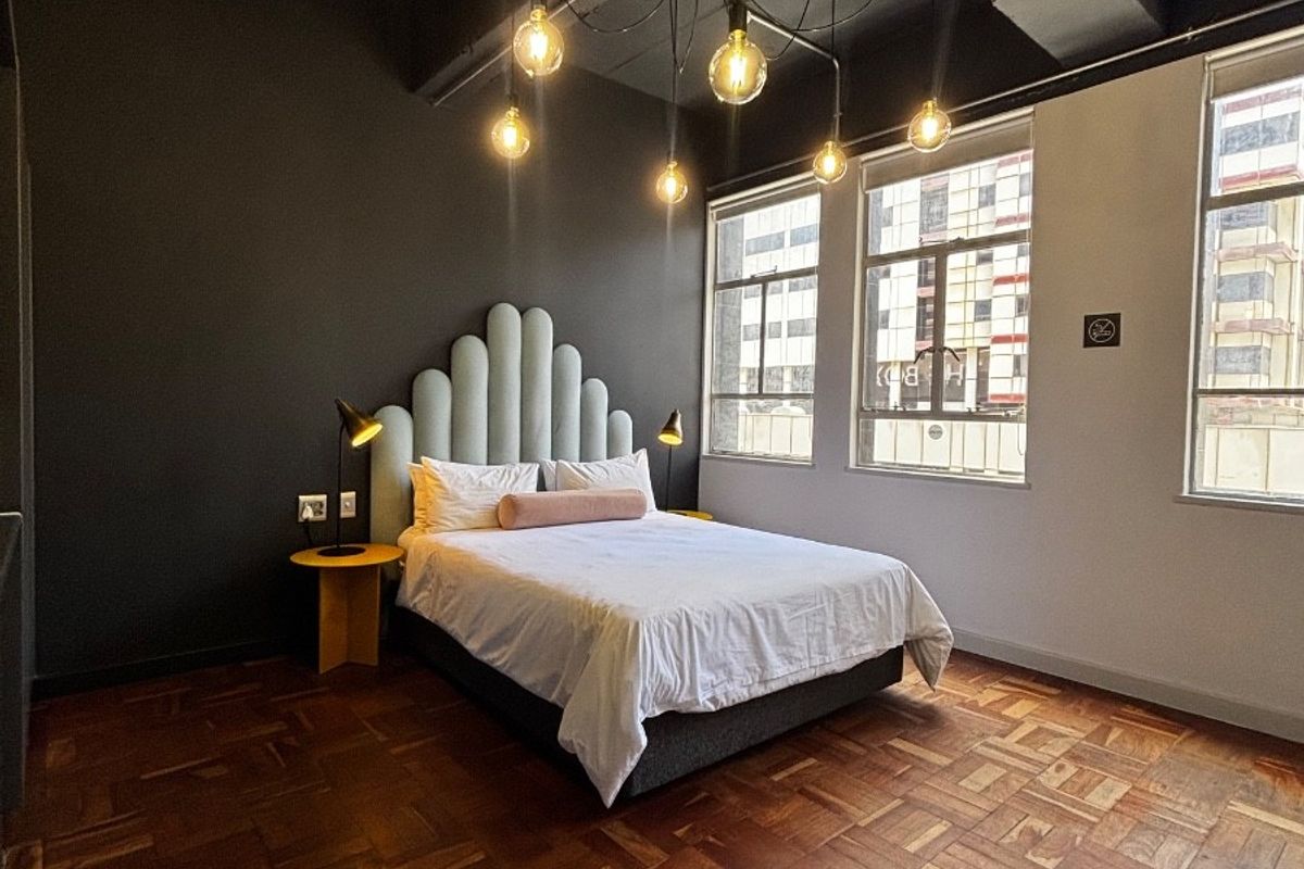 A stylish bedroom featuring original parquet flooring, exposed concrete ceilings, and industrial-style pendant lighting, complemented by large windows providing ample natural light.