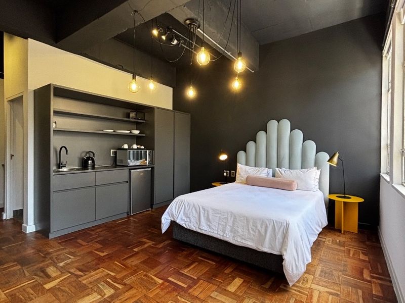 A modern open-plan studio apartment featuring dark industrial-style ceilings, rich parquet flooring, and a compact built-in kitchenette area.