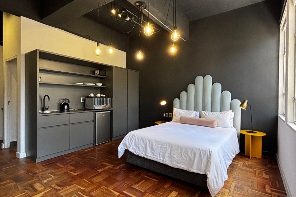 A modern open-plan studio apartment featuring dark industrial-style ceilings, rich parquet flooring, and a compact built-in kitchenette area.