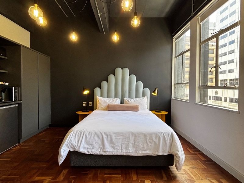 A modern bedroom featuring dark walls, exposed ceiling details, warm parquet flooring, and large windows providing ample natural light.