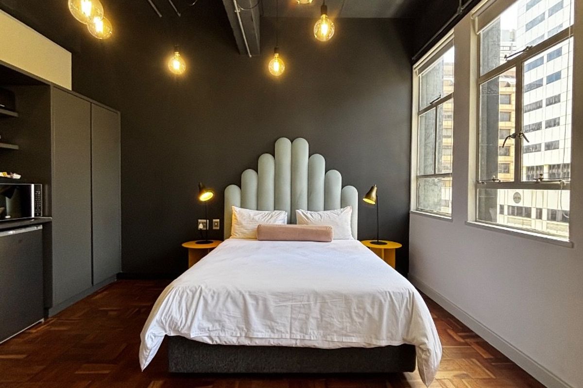 A modern bedroom featuring dark walls, exposed ceiling details, warm parquet flooring, and large windows providing ample natural light.
