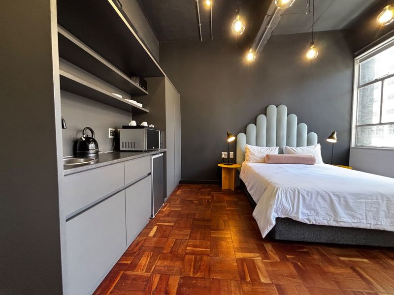 A modern open-plan studio apartment featuring dark cabinetry, a kitchenette, parquet flooring, and an industrial-style exposed ceiling with large windows.