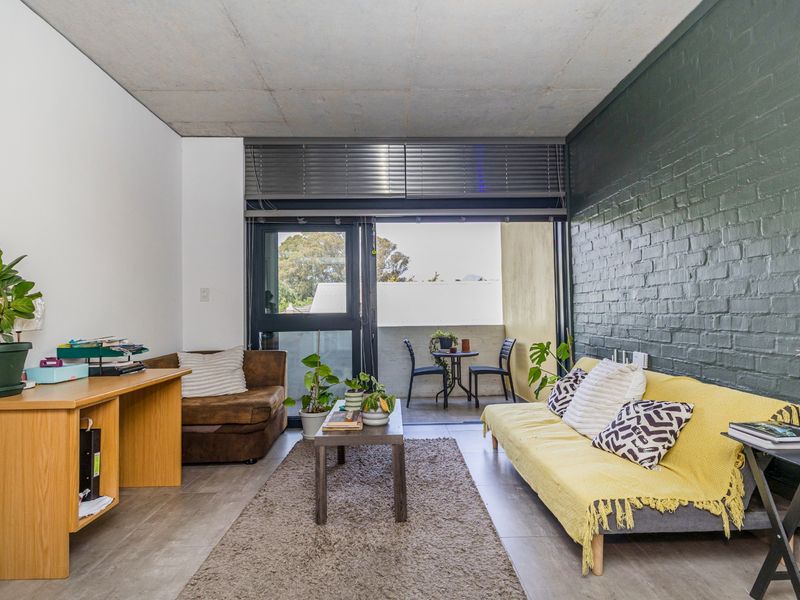 A modern living room featuring an exposed concrete ceiling, a dark painted brick feature wall, and large sliding doors leading to a private balcony.