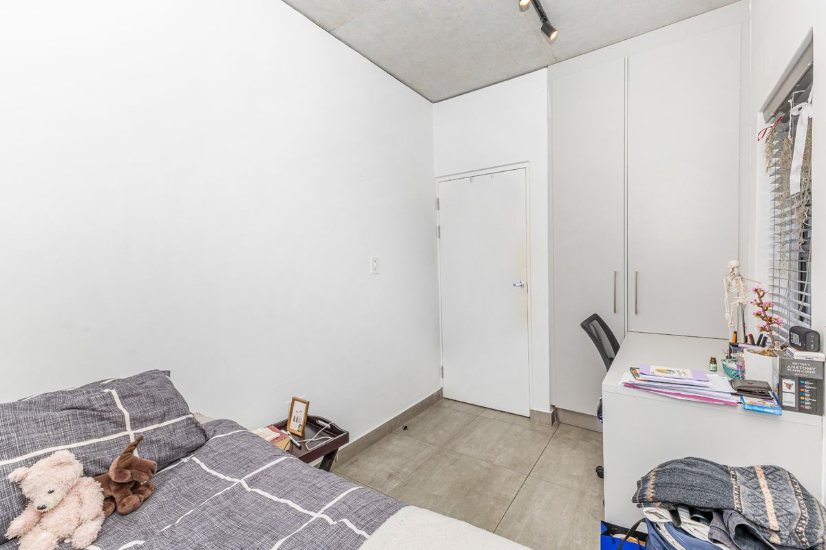 A compact bedroom featuring tiled flooring, an exposed concrete ceiling, and built-in cupboards with integrated desk space.
