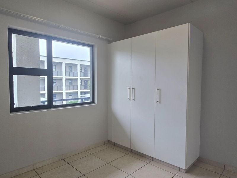 A clean, empty bedroom featuring neutral tiled flooring, a built-in white wardrobe, and a large window providing natural light.