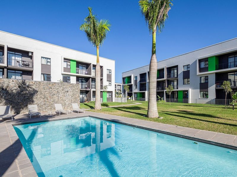 A well-maintained communal swimming pool area featuring a stone feature wall, paved decking, and landscaped lawn within a modern apartment complex.