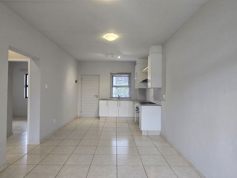 An open-plan living and kitchen area featuring neutral tiled flooring, clean white cabinetry, and integrated cooking appliances in a bright, vacant space.