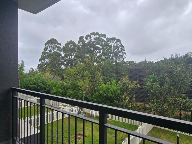 A private balcony featuring a modern metal balustrade, overlooking a landscaped garden area with mature trees and a white picket fence.