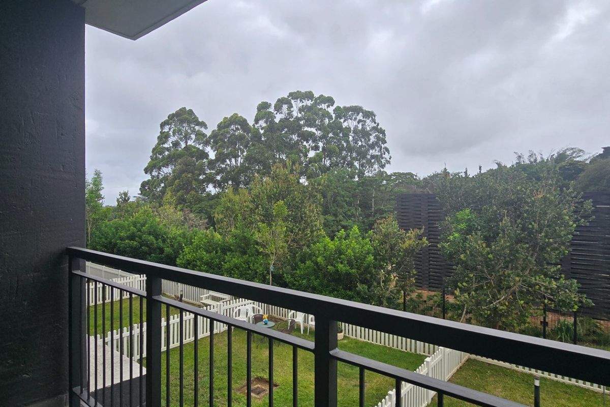 A private balcony featuring a modern metal balustrade, overlooking a landscaped garden area with mature trees and a white picket fence.
