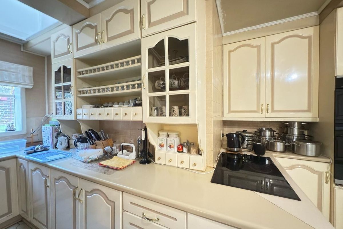 A functional kitchen featuring traditional cream cabinetry with glass-fronted display units, a ceramic hob, and tiled splashbacks, offering a classic aesthetic with potential for modernization.