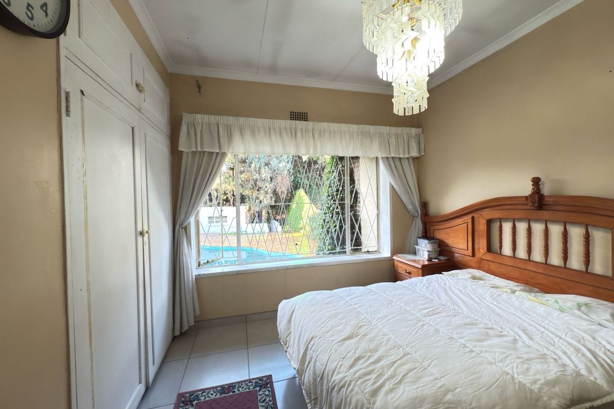 A bedroom featuring built-in cupboards, tiled flooring, and security bars on the window, offering a view of the garden and pool area.
