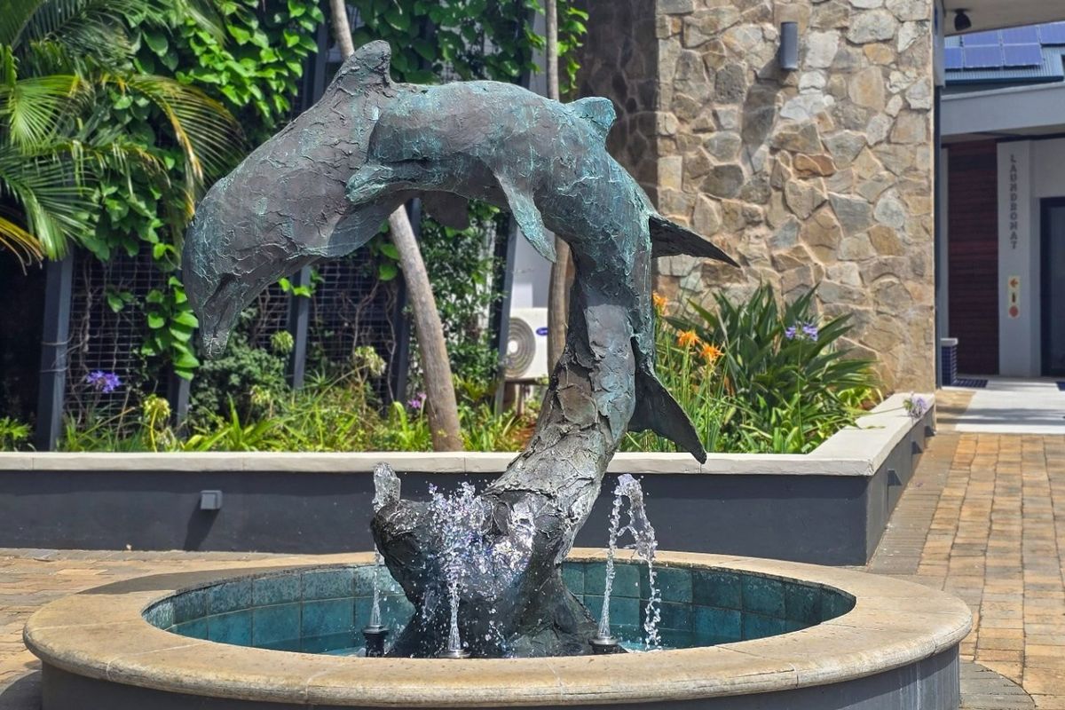 A well-maintained outdoor courtyard featuring a decorative dolphin fountain, stone-clad wall accents, and paved walkways within a landscaped garden setting.