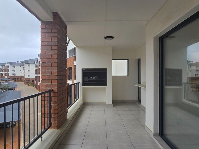 A spacious covered balcony featuring a built-in braai, tiled flooring, and a metal balustrade, offering views of the apartment complex.
