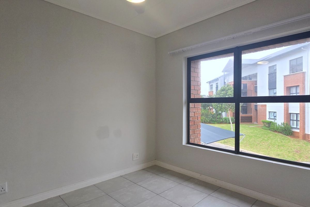 A bright, empty bedroom featuring neutral tiled flooring, clean walls, and a large window overlooking the apartment complex grounds.