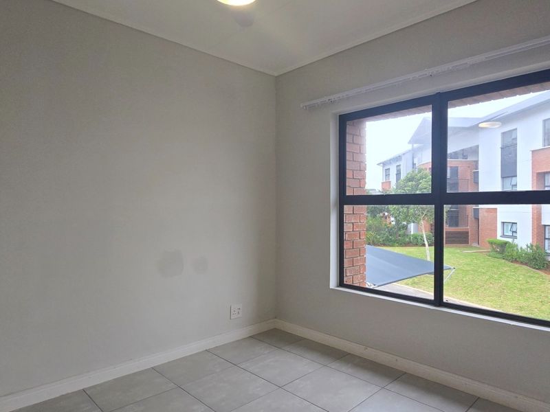 A bright, empty bedroom featuring neutral tiled flooring, clean walls, and a large window overlooking the apartment complex grounds.